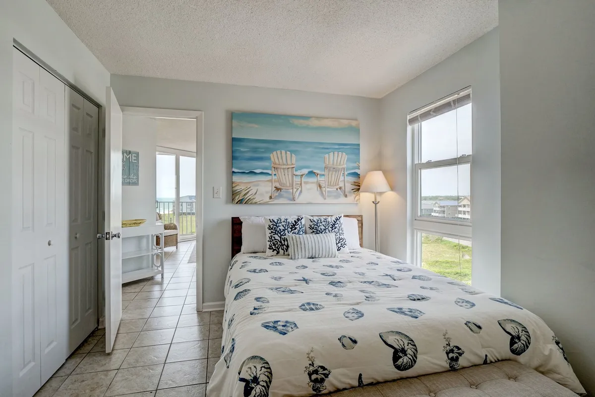 This image features a serene bedroom decorated with beach-themed artwork, a queen-sized bed with seashell-patterned bedding, a lamp, and a window offering a soothing view. The tranquil setting is designed for rest and relaxation, with natural light filtering into the space.