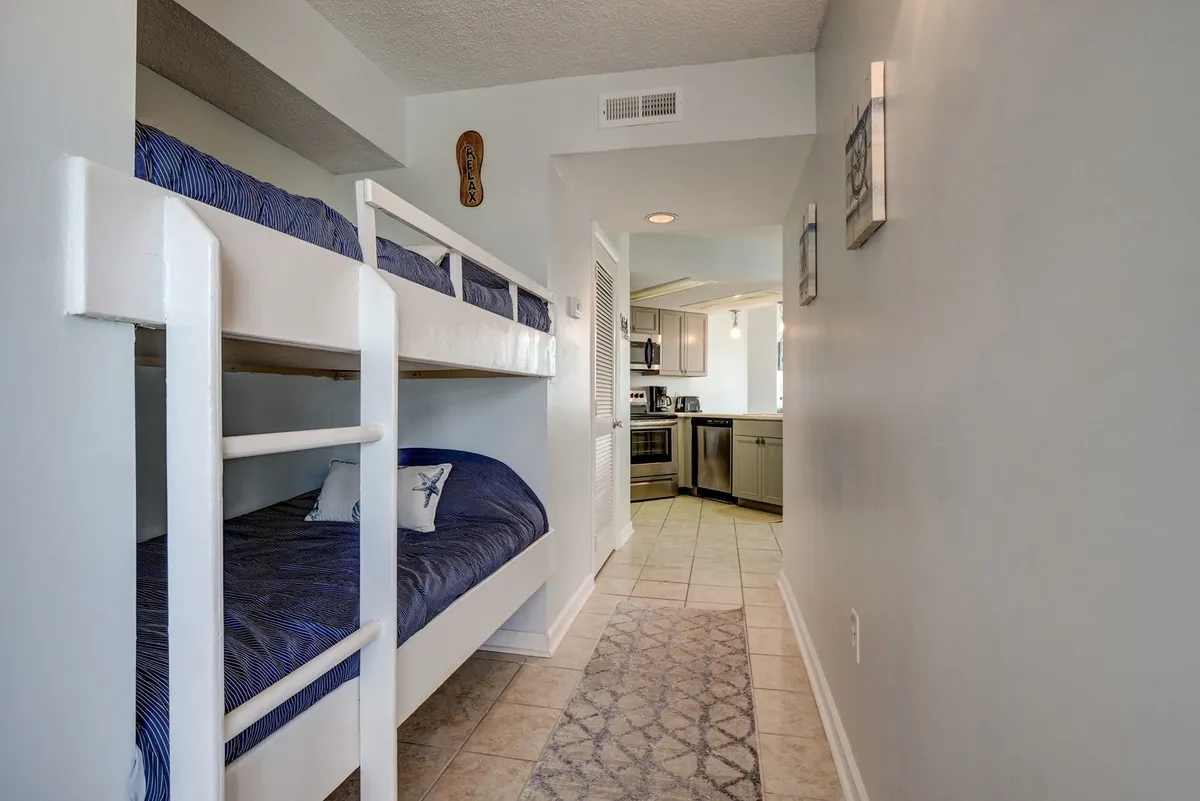 This image shows a built-in bunk bed nook situated along a hallway, complete with cozy bedding and a textured rug. In the background, part of the kitchen area is visible, blending the nook into the main living space. The scene is warm and family-friendly, suggesting a space where kids can relax, read, or nap while staying close to the action of the home.