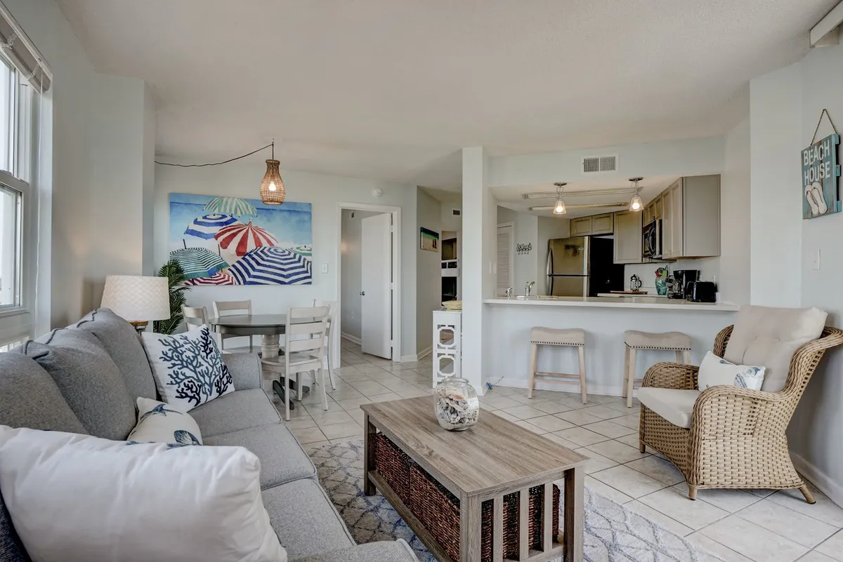 An open-concept living and dining area featuring a plush sofa with decorative pillows, a coffee table adorned with beach-themed décor, and a dining table set for shared meals. Natural light streams in from windows, and comfortable seating encourages relaxation and socializing.