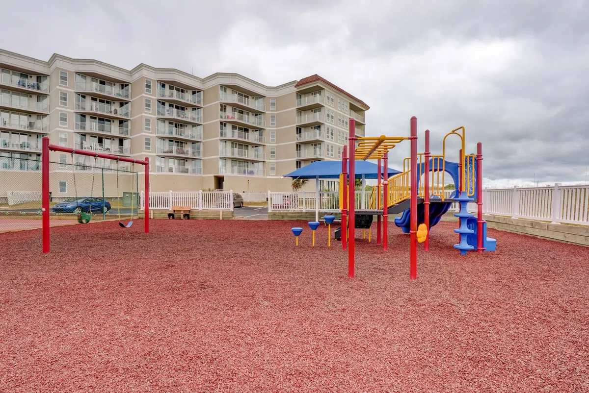 A vibrant playground area with modern equipment and a swing set, set against the backdrop of an apartment building. Children are seen playing, and families gather nearby, suggesting a joyful, community-oriented atmosphere. The lush grass and open space make it perfect for young kids to run around and socialize.