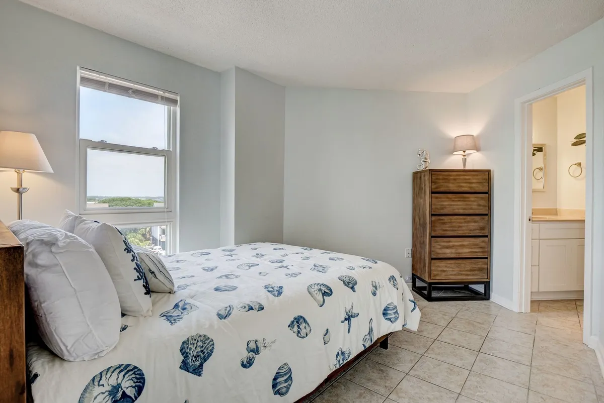 A peaceful bedroom featuring a cozy bed with a shell-patterned duvet, a wooden dresser, lamp, and a window filling the space with natural daylight. The setting offers a soft, inviting atmosphere ideal for unwinding and enjoying quiet moments.