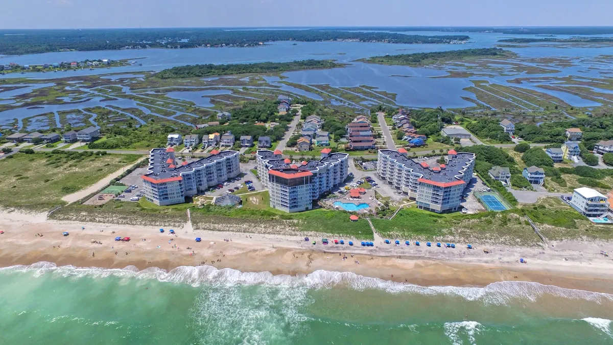 A scenic exterior showing beachfront condominiums with a large swimming pool in the foreground, golden sand and surf beyond, and lush coastal vegetation and marshlands. The image captures a lively yet peaceful atmosphere ideal for beach activities and family gatherings.