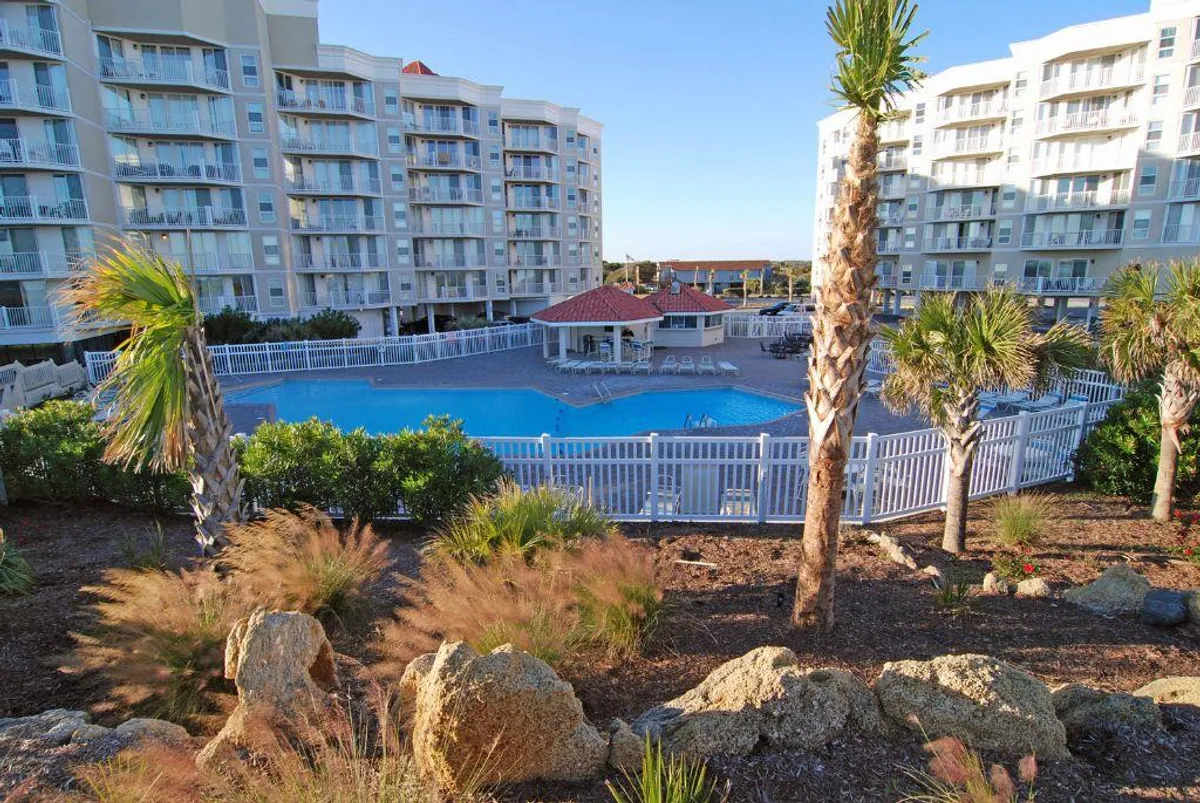 An inviting view of the resort's exterior swimming pool area, complete with palm trees, rocky landscaping, and the building's facade in the background. People are seen swimming, sunbathing, and socializing, all bathed in the warm glow of sunset, with barbecue spaces nearby.
