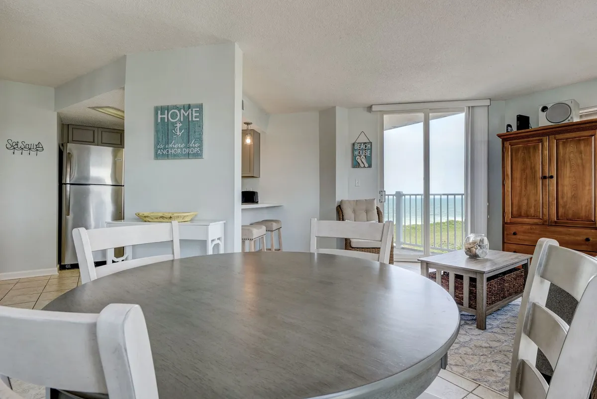 A bright, inviting interior scene featuring a round dining table with beach-themed decor and a large sliding glass door that opens to a sweeping view. Natural light pours in, suggesting a seamless connection between the indoor dining area and the outdoor scenery, ideal for cozy meals or morning coffee with an ocean backdrop.