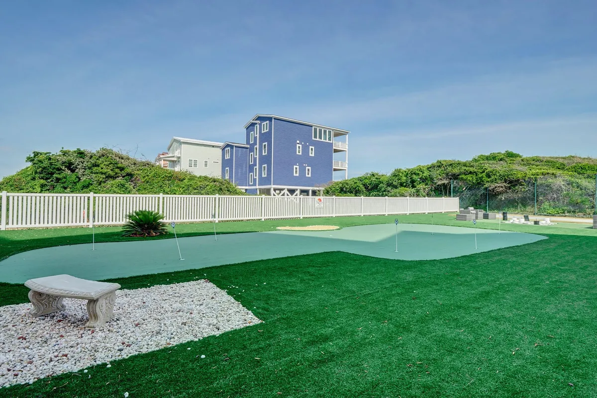 A peaceful, green putting green with a stone bench and a white fence, overlooking a blue house in the background. The area is designed for relaxation and casual outdoor gatherings, with plenty of sunlight and open space.