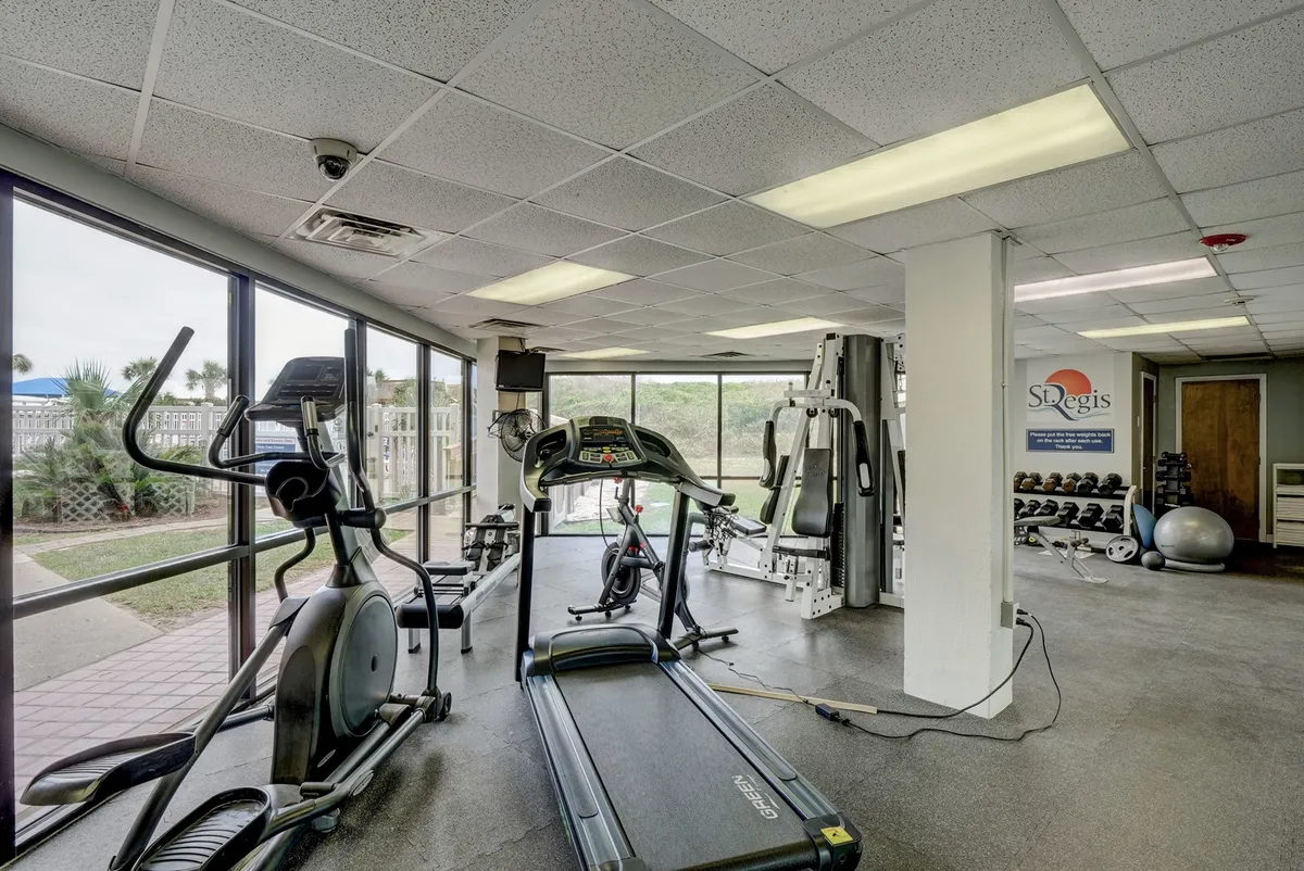 A modern fitness center interior with treadmills, elliptical machines, and weightlifting equipment. Sunlight shines through wide windows, offering a glimpse outside and creating an energizing atmosphere for wellness routines.
