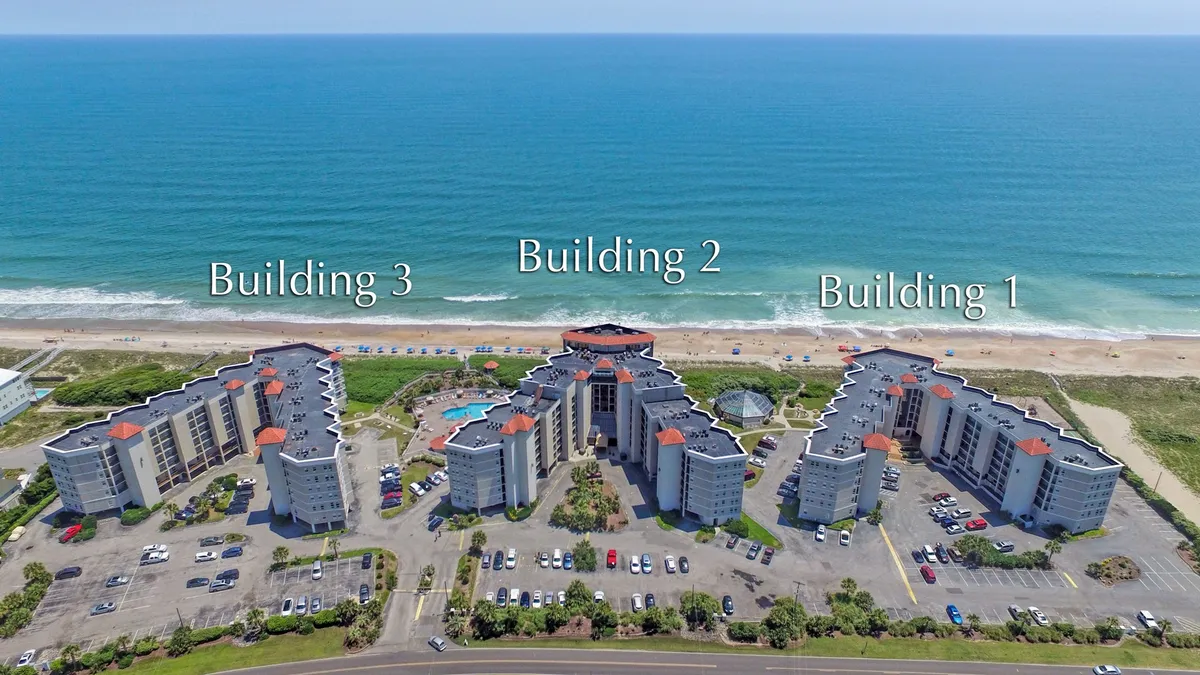 A wide exterior shot of three condominium buildings overlooking a sparkling swimming pool, with the ocean visible in the background. Lounge chairs and umbrellas surround the pool, inviting families to relax and cool off after a day at the beach.