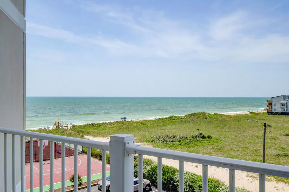 The image offers a sweeping view from the condo's balcony, overlooking the sparkling Atlantic Ocean, a lush green lawn, a basketball court, and distant houses along North Topsail Beach. The serene setting captures the promise of sunrise over the waves and space for outdoor activities below.
